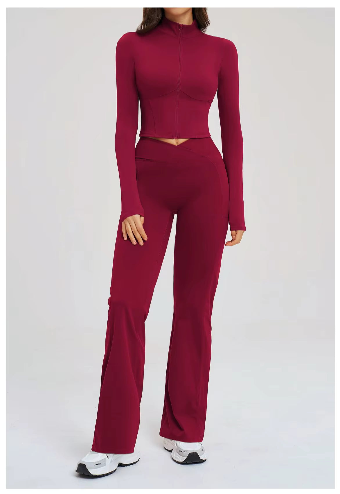 Woman wearing a fitted burgundy two-piece athleisure set consisting of a long-sleeve cropped top and high-waisted flared leggings, paired with white sneakers, standing against a neutral background.
