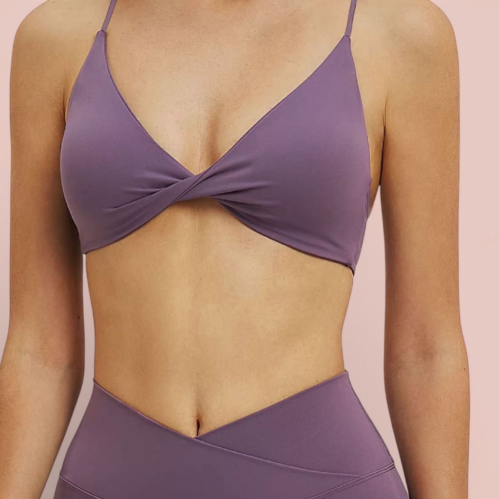 Woman wearing a mauve sports bra with thin straps and a twisted front detail, paired with high-waisted matching leggings, shown from shoulders to hips against a soft neutral background.
