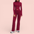 Woman wearing a fitted burgundy athleisure set with a long-sleeve cropped top and high-waisted flared leggings, paired with white sneakers, standing against a soft beige background.