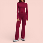 Woman wearing a fitted burgundy athleisure set with a long-sleeve cropped top and high-waisted flared leggings, paired with white sneakers, standing against a soft beige background.