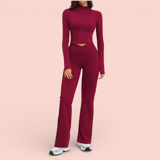Woman wearing a fitted burgundy athleisure set with a long-sleeve cropped top and high-waisted flared leggings, paired with white sneakers, standing against a soft beige background.
